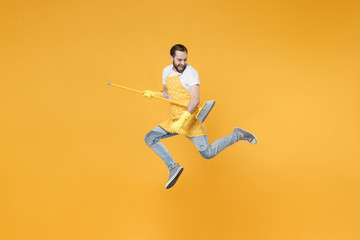 Cheerful young man househusband in apron rubber gloves hold broom like guitar while doing housework isolated on yellow wall background studio portrait. Housekeeping concept. Jumping, having fun.