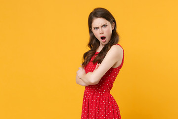 Side view of shocked irritated young brunette woman girl in red summer dress posing isolated on yellow background. People lifestyle concept. Mock up copy space. Holding hands crossed, looking camera.