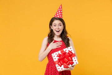 Joyful young woman girl in red summer dress birthday hat isolated on yellow background. Birthday holiday concept. Mock up copy space. Hold white red present box with gift ribbon bow put hand on chest.