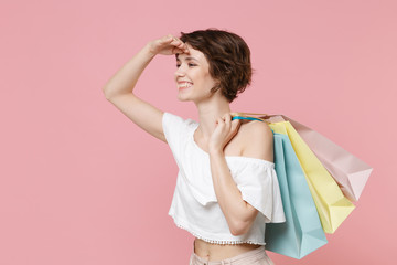 Side view of smiling young woman girl in summer clothes hold package bag with purchases isolated on pink background. Shopping discount sale concept. Holding hand at forehead looking far away distance.