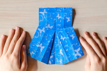 Girl's hands making origami blue dress from butterfly printed paper. Step by step instruction, step14. Mother's day, birhday, summer gift, family leisure time concept. DIY tutorial, flatlay, top view