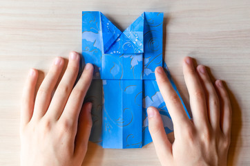 Girl's hands making origami blue dress from butterfly printed paper. Step by step instruction, step15. Mother's day, birhday, summer gift, family leisure time concept. DIY tutorial, flatlay, top view