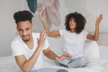 Fototapeta premium Young angry sad irritated frustrated african swearing screaming couple girl guy in white t-shirts sitting on bed indoors in bedroom at home spending time in room. Bad mood quarantine lifestyle concept
