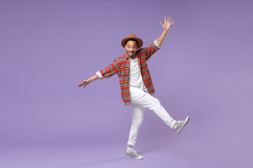 Fototapeta na wymiar Excited young african american guy in casual colorful shirt hat posing isolated on violet background studio portrait. People lifestyle concept. Mock up copy space. Dancing rising leg, spreading hands.