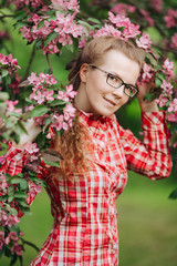 Beautiful young happy woman with glasses in a flowered garden. Girl among the flowers of apple trees, cherries. Springtime scene. Positive model in a casual style. Lifestyle