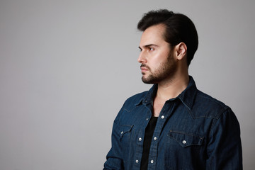 Obraz premium Side view of bearded man in the denim outfit which mixed with bright white t-shirt posing over white wall.