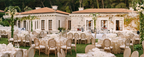 Wedding dinner table reception. Elegant tables for guests with cream tablecloths with patterns, on green lawn, with garlands and chandeliers hanging over them. Chairs with round back - Powered by Adobe