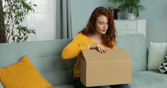 Young Caucasian pretty woman sitting on couch in living room and opening carton box. Red-haired attractive cheerful girl getting parcel at home. Mail delivery to house.