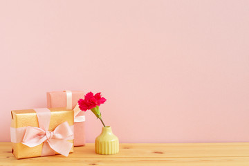 Gift boxes with pink spray carnation flower in vase on wooden table with pink background. Anniversary concept, floral arrangement, copy space