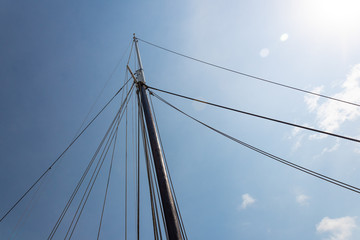 Mast of a vintage sailing vessel with numerous rigging lines coming from the top in many directions, against a bright blue sky with clouds, horizontal aspect