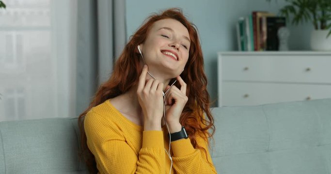 Close up of red-haired Caucasian young pretty joyful woman in headphones sitting on sofa at home. Beautiful girl listening to music on smartphone, singing and smiling. Female enjoying song.