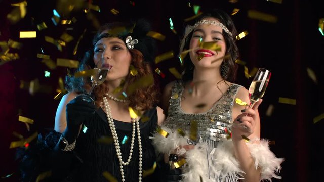 Beautiful flappers women dressed in Roaring twenties time drinking champagne under confetti rain. Vintage, retro party, fashion, girls friends concept