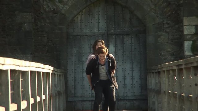 Happy young smiling couple playfully running piggyback along wooden castle bridge on warm sunny day low angle centered long shot slow motion