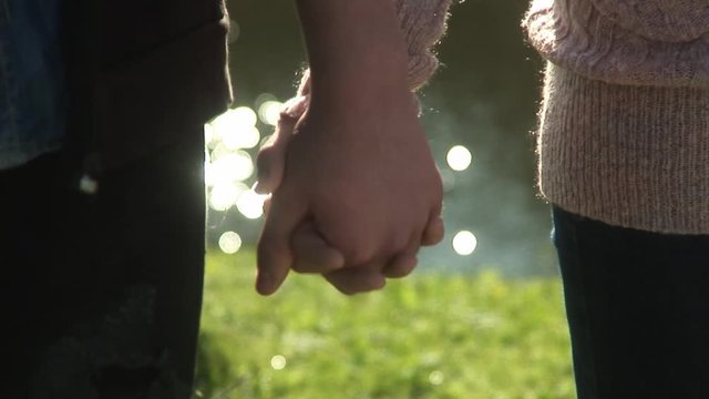 Couple holding hands standing along picturesque lake during warm summers day at park lake handheld close up shot