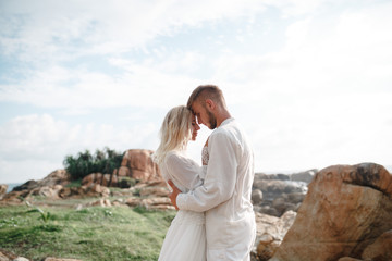 Fototapeta premium Beautifull wedding couple kissing and embracing near the shore of indian ocean standing on sea cliff. Close up happy young wedding couple with sea on the mountain