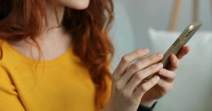 Close up of smartphone in hands of Caucasian woman with red curly hair. Female tapping and scrolling on mobile phone. At home. Indoor. Girl texting message on cellphone device. Chatting online.