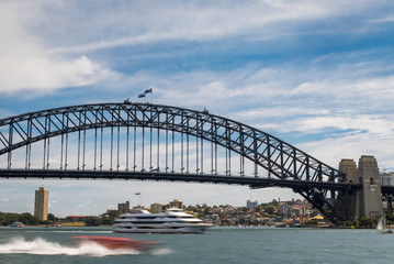A ferry boat passing under the Sydney Harbour Bridge structure with cloudy blue sky above and city buildings in the background.