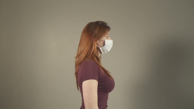 Young attractive girl putting on surgical Mask for corona virus, caucasian woman, standing and facing at camera in studio, during covid-19 pandemic in France. One person profile Medium shot.