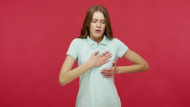 Heart attack. Stressed young woman in polo t-shirt clutching chest, needs emergency of acute myocardial infarction, difficult to breath, risk of asthma. indoor studio shot isolated on red background