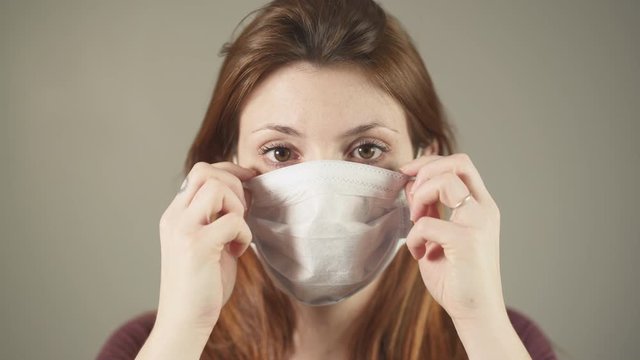 Young girl putting on surgical Mask for corona virus prevention, caucasian woman, red hair and shirt standing and facing at camera in studio, during covid-19 pandemic in France. One person close-up.