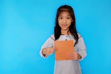 A cute young Asian girl standing against a plain light blue background, holding a blank piece of paper, smiling.
