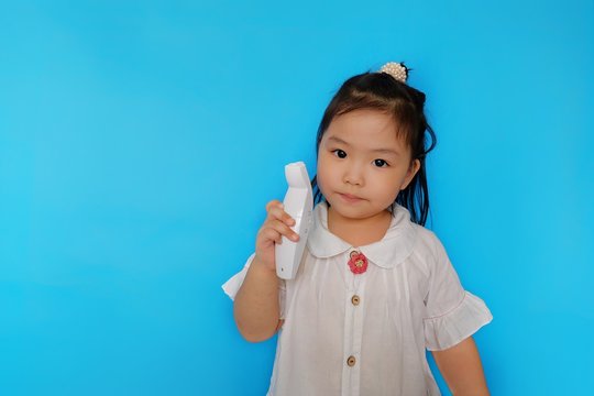 A Cute Young Asian Girl Is Role Playing As A Doctor, Holding Up An Infrared Thermometer Trying To Check The Temperature Of Her Family Members, Looking Serious. Plain Light Blue Background.