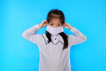A cute Asian girl putting on a white cloth mask, covering her mouth and nose before going out to protect against Covid-19, with plain light blue background.