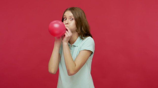 Funny happy young brunette woman in polo t-shirt inflating balloon with great effort, laughing and dancing, getting ready for party, celebrating birthday. indoor studio shot isolated on red background