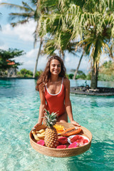Pretty smiling young woman in red swimsuit is preparing to eat breakfast in the swimming pool. Girl...