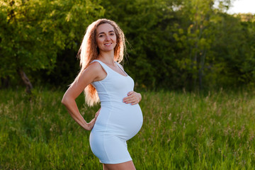Naklejka na ściany i meble Beautiful smiling pregnant woman with a good figure in a slinky white summer dress on a natural background