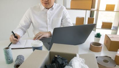 Young asian man looking on computer laptop and taking writing order list on paper with business owner concept.