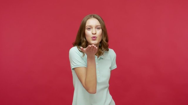 Love you all! Playful charming young brunette woman in polo t-shirt sending air kisses around, flirting and demonstrating romantic feelings, affection. indoor studio shot isolated on red background