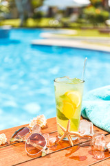 Glass of cclassic mojito cocktail and caipirinha with shells, sunglasses and blue towel closed to swimming pool. Vacation, summer, holiday, luxury resort concept.
