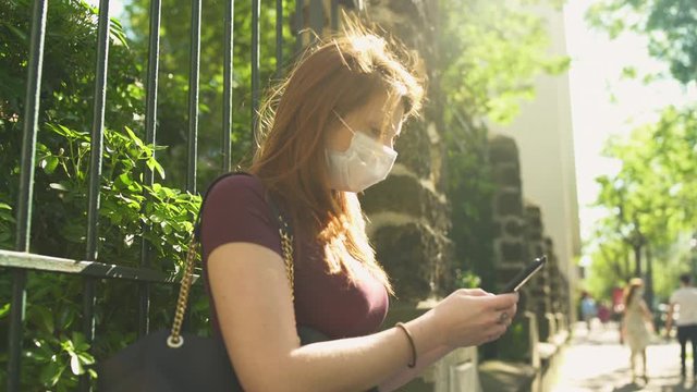 Cute woman with surgical mask, redhead and freckles, red hair and shirt sitting outside in street, during covid-19 pandemic in France. Sending text messages with smartphone. 4K UHD.
