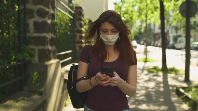 Caucasian woman with surgical mask, redhead and freckles, red hair and shirt walking outside in street, during covid-19 pandemic in France. Sending text messages with smartphone. Phone conversation.