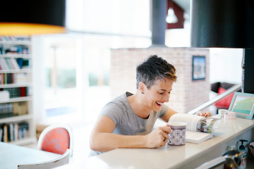 Short-haired woman with a cup and a book inside the house and laughing