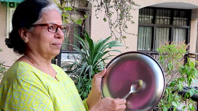 Senior citizen Indian woman beating a place with spoon as a mark of respect for essential healthcare workers, medical personel, doctors, policemen and more during the coronavirus lockdown in India