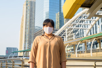 Overweight Asian woman with mask at skywalk bridge against view of the city