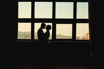 Fototapeta na wymiar dark silhouette of a loving couple, man and woman no faces, against of a huge stained glass window with a view of the city and curtains on the sides. The comfort of home, a life together for couples