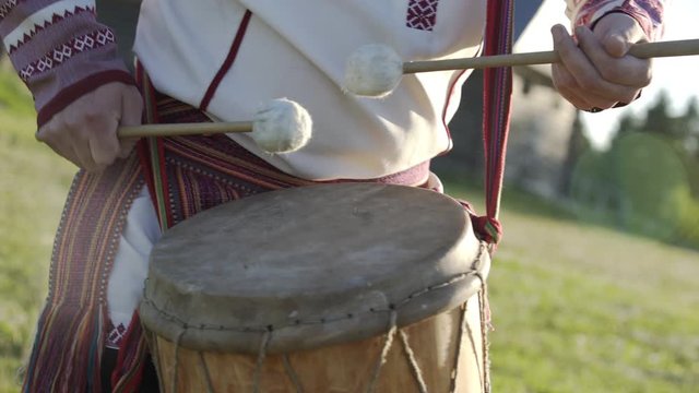 Man in traditional russian clothes dancing and playing on folklore drum outdoor on traditional antique wooden windmill background. Man wearing national Finno-Ugric clothes.