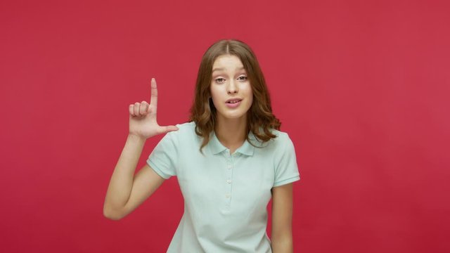 You lost, your fault! Young brunette woman in polo t-shirt making loser gesture and pointing to camera, teasing and blaming for mistake, unsuccess. indoor studio shot isolated on red background
