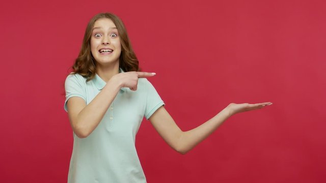 Overjoyed enthusiastic girl holding copy space on palm and looking excitedly, pointing at workspace mockup for advertisement, empty place for promotional image. studio shot isolated on red background