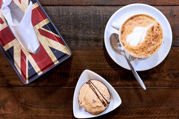 Cappuccino in a white cup on a plate with a spoon. The cookie in a small triangular saucer. English Flag Napkin Box on a wooden table. View from above