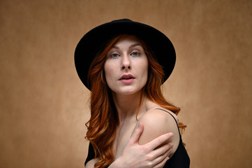 Close-up portrait of a young pretty woman with long red hair on a beige background. Model posing in a black hat.