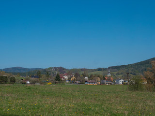 Spring landscape with view on village Cvikov in Lusitian mountains with old and modern houses and lush green grass meadow, deciduous and spruce tree forest and hills, blue sky background