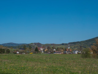 Spring landscape with view on village Cvikov in Lusitian mountains with old and modern houses and lush green grass meadow, deciduous and spruce tree forest and hills, blue sky background