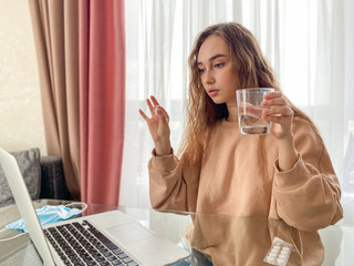 Sad, upset, young woman holding a glass of water and pills sitting in the living room, feeling ill.