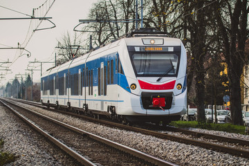 White passenger train with blue and red details running on double train tracks from Trieste centrale towards Ljubljana, Slovenia on a gray dull autumn afternoon.