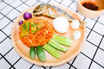 Fried rice with prik pao (Chilli paste with soya bean oil), omelet with cha om, boiled chicken egg, cucumber, eggplant and fried mackerel in wooden plate put on check pixel plaid black and white cloth