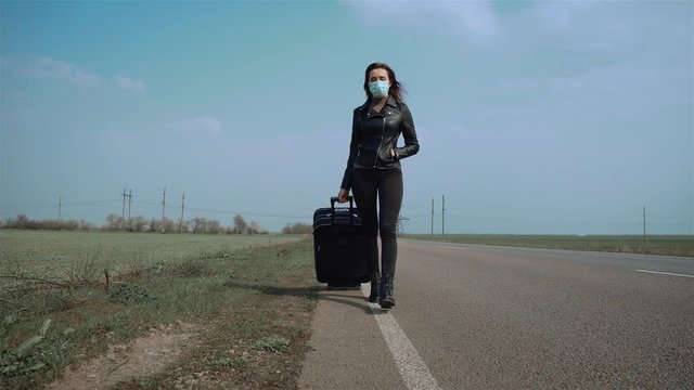 Young caucasian woman in protective face mask and black jacket walking across road for stopping car with suitcase on wheels. Dangerous COVID-19 infection